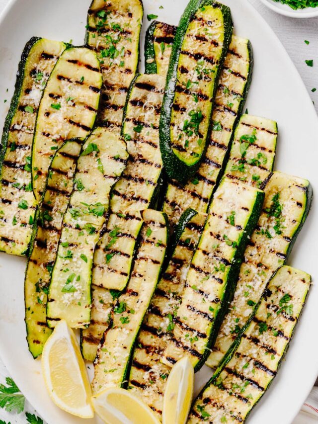 Overhead view of grilled zucchini arranged on a white serving platter and topped with grated Parmesan cheese and chopped fresh parsley. Lemon wedges are tucked alongside the zucchini, with crisp grill marks visible on every slice.