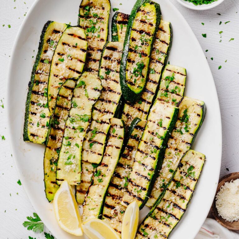 Overhead view of grilled zucchini arranged on a white serving platter and topped with grated Parmesan cheese and chopped fresh parsley. Lemon wedges are tucked alongside the zucchini, with crisp grill marks visible on every slice.