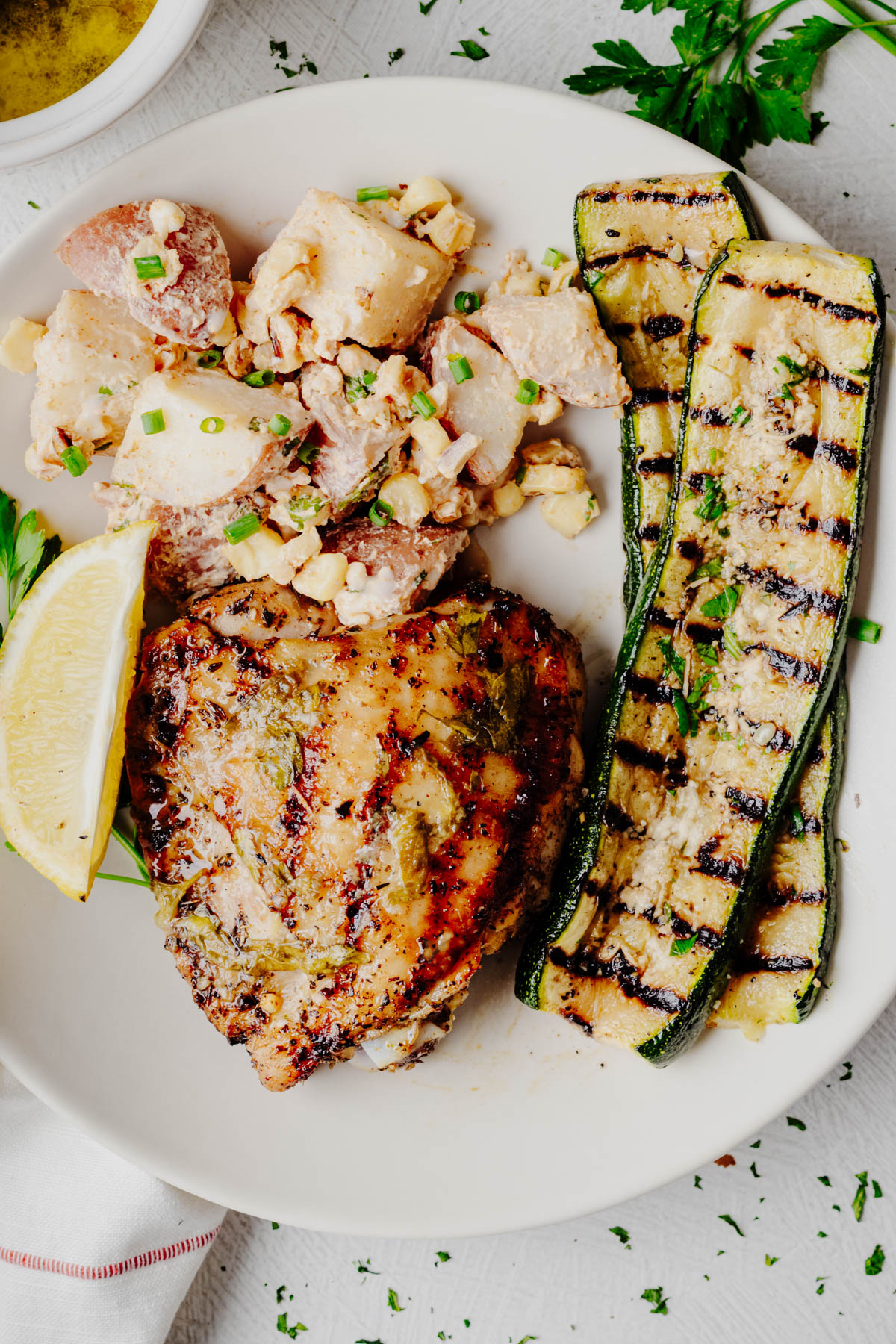 A dinner plate with grilled lemon herb chicken thighs, creamy potato salad, and grilled zucchini topped with grated Parmesan, fresh parsley, and served with a lemon wedge.