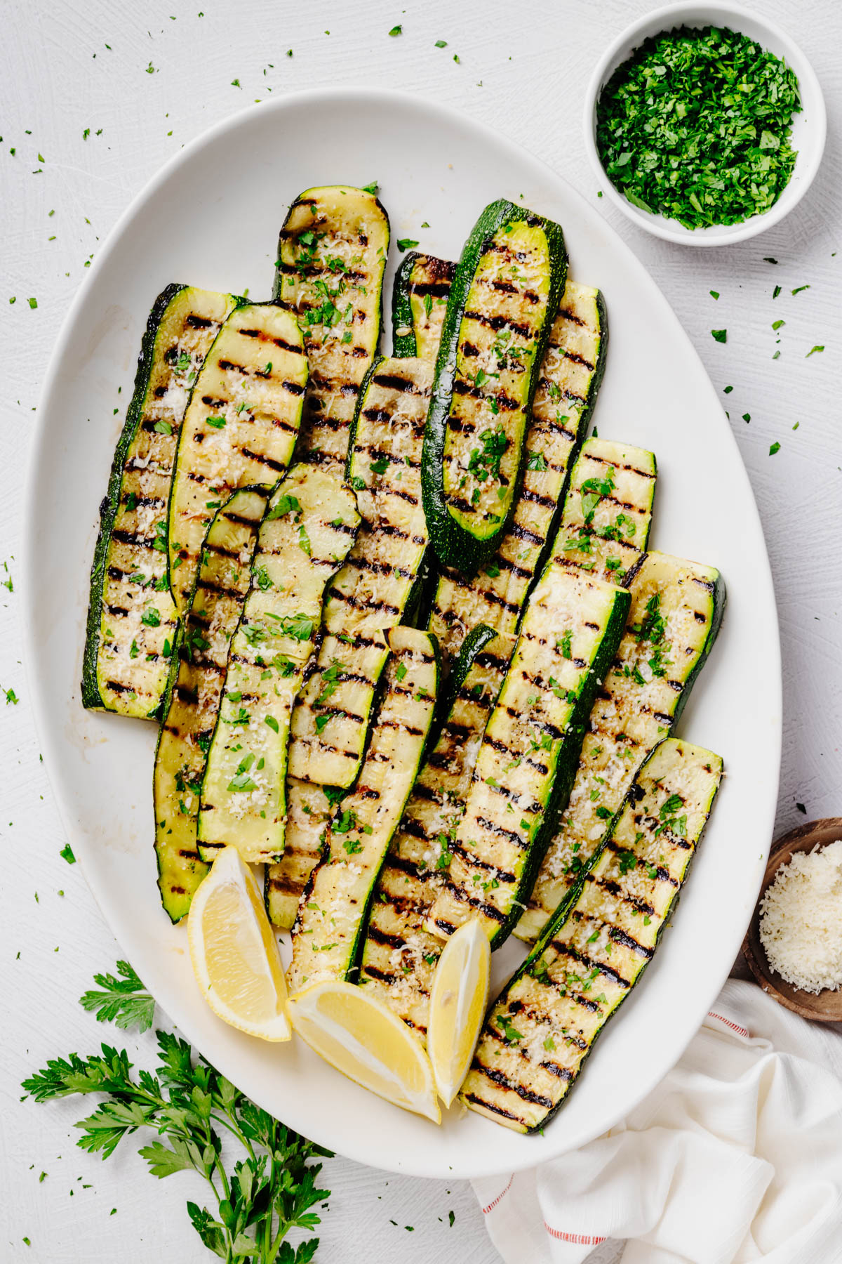 Grilled zucchini arranged on a white serving platter with fresh parsley, grated Parmesan cheese, and lemon wedges. The zucchini is topped with grated Parmesan and chopped parsley and has dark grill marks throughout.