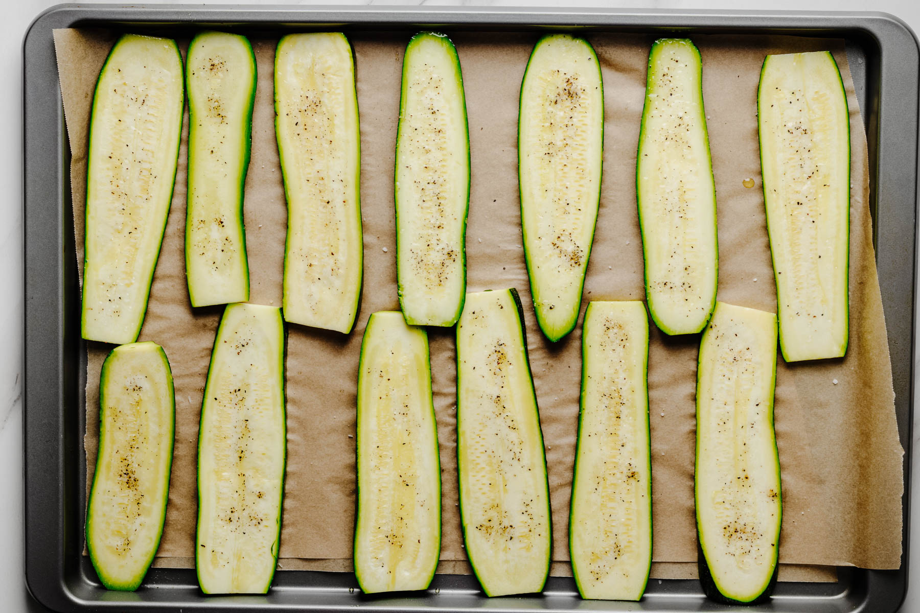 Fresh zucchini planks arranged in a single layer on a parchment-lined baking sheet after being sliced and seasoned.