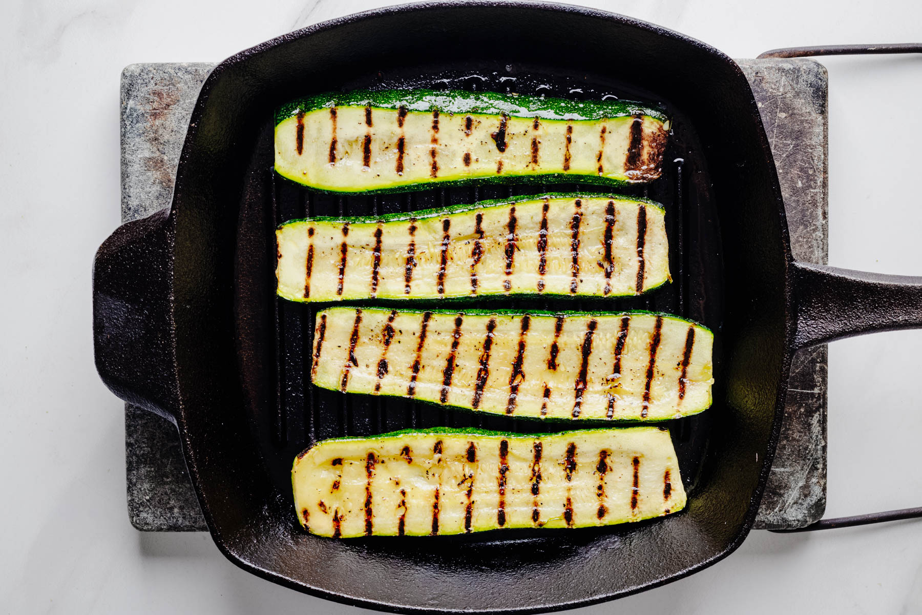 Four zucchini planks cooking on a cast iron grill pan with dark grill marks beginning to develop.