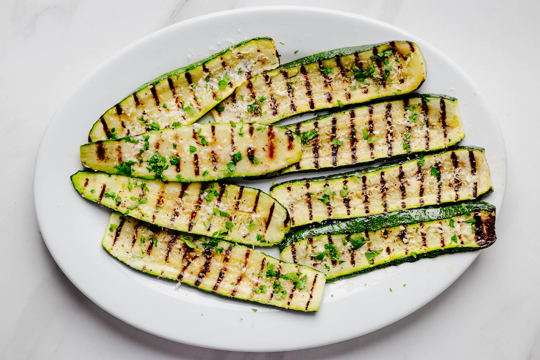 Grilled zucchini arranged on a white serving platter and topped with grated Parmesan cheese and chopped fresh parsley before serving. This image shows the final step for making homemade Parmesan grilled zucchini from Heavenly Home Cooking.