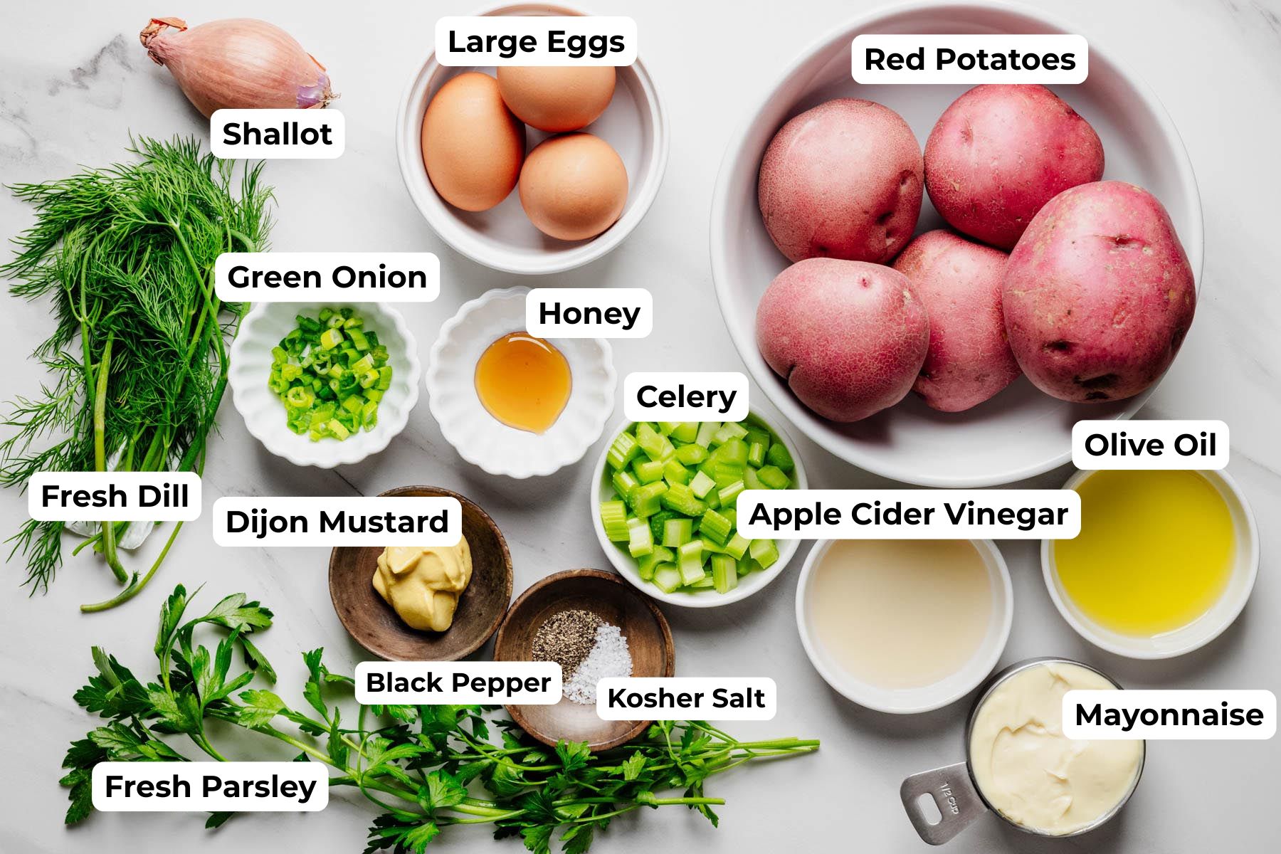 Labeled ingredients for red potato salad arranged on a marble surface, including red potatoes, eggs, mayonnaise, olive oil, apple cider vinegar, Dijon mustard, honey, celery, green onion, shallot, fresh dill, fresh parsley, kosher salt, and black pepper.