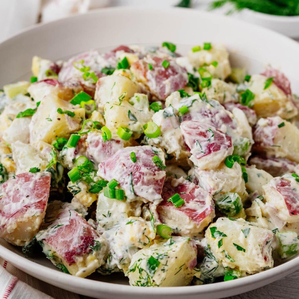 Close-up of creamy red potato salad made with red potatoes, hard-boiled eggs, fresh dill, celery, and green onions in a white serving bowl.