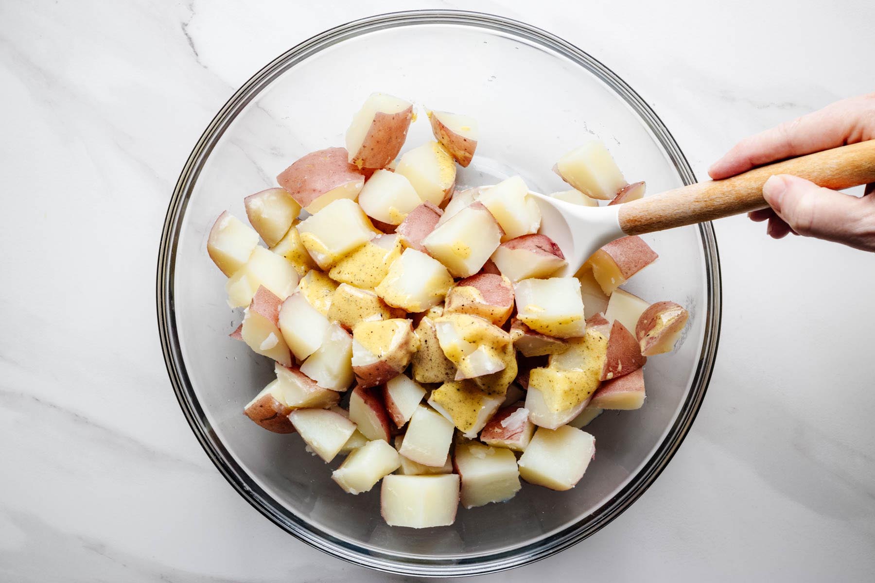 Warm red potatoes being tossed with Dijon vinaigrette in a mixing bowl.