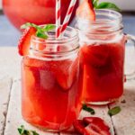 Two mason jar mugs filled with strawberry basil agua fresca sit on a light stone surface, garnished with fresh strawberry slices and basil leaves. Red-and-white striped paper straws add a festive touch, while a pitcher of the drink is softly blurred in the background.