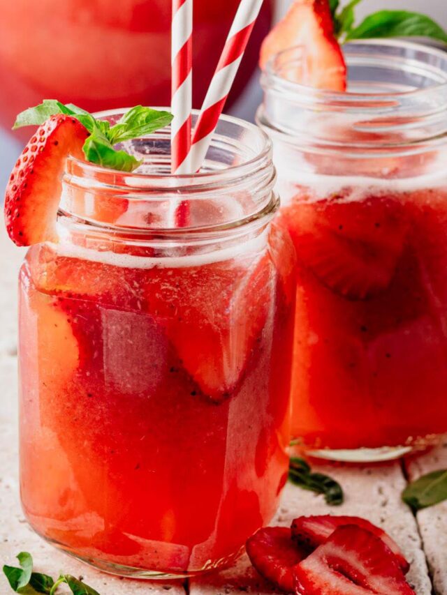 Two mason jar mugs filled with strawberry basil agua fresca sit on a light stone surface, garnished with fresh strawberry slices and basil leaves. Red-and-white striped paper straws add a festive touch, while a pitcher of the drink is softly blurred in the background.