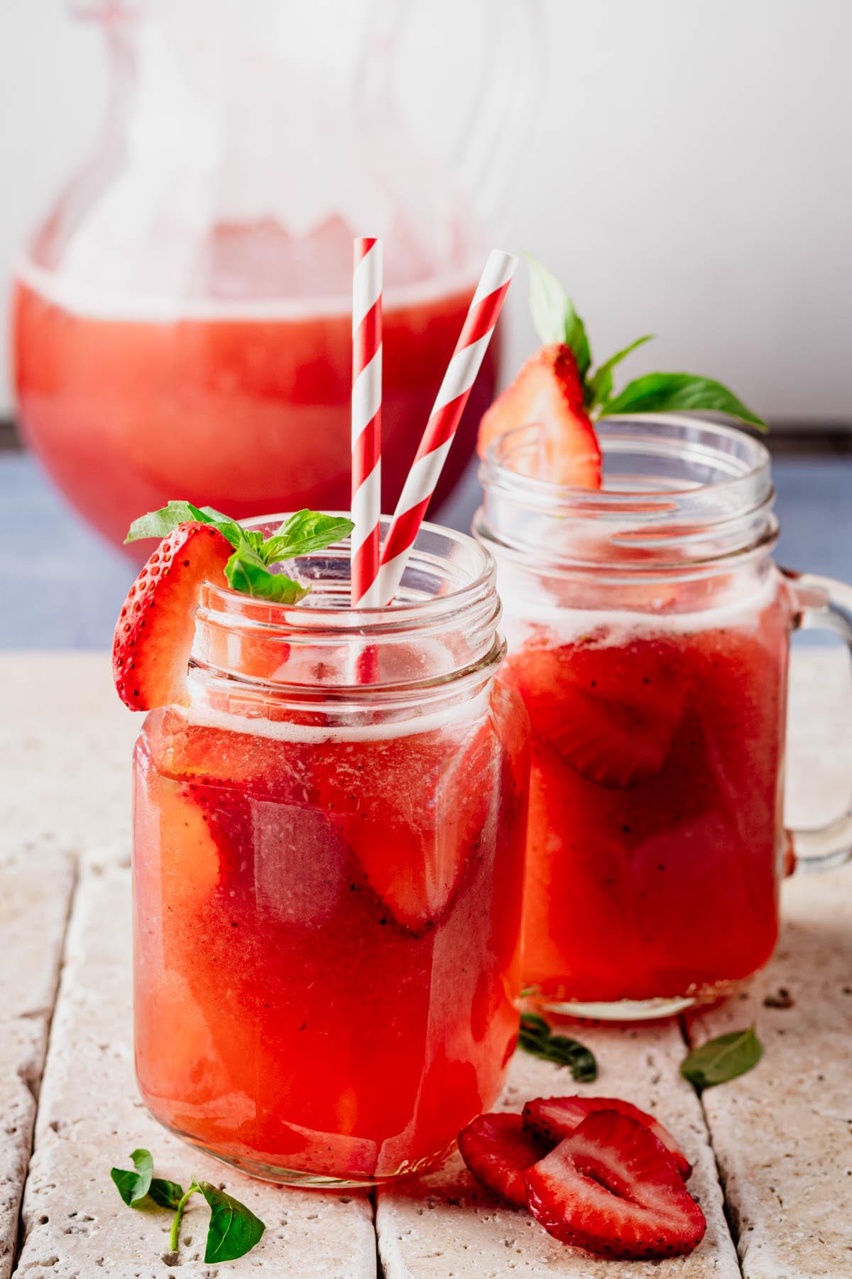 Mason jar mugs filled with strawberry agua fresca garnished with fresh strawberries and basil, with a pitcher of agua fresca in the background.