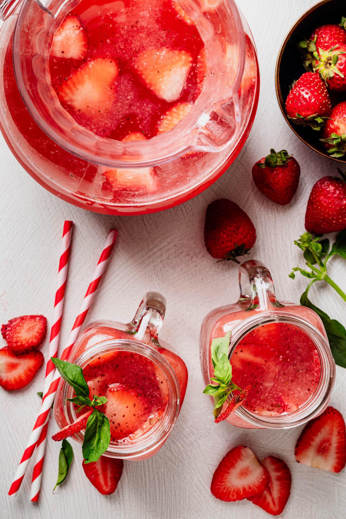 Overhead view of strawberry agua fresca in mason jar mugs beside a pitcher, fresh strawberries, basil, and striped paper straws.