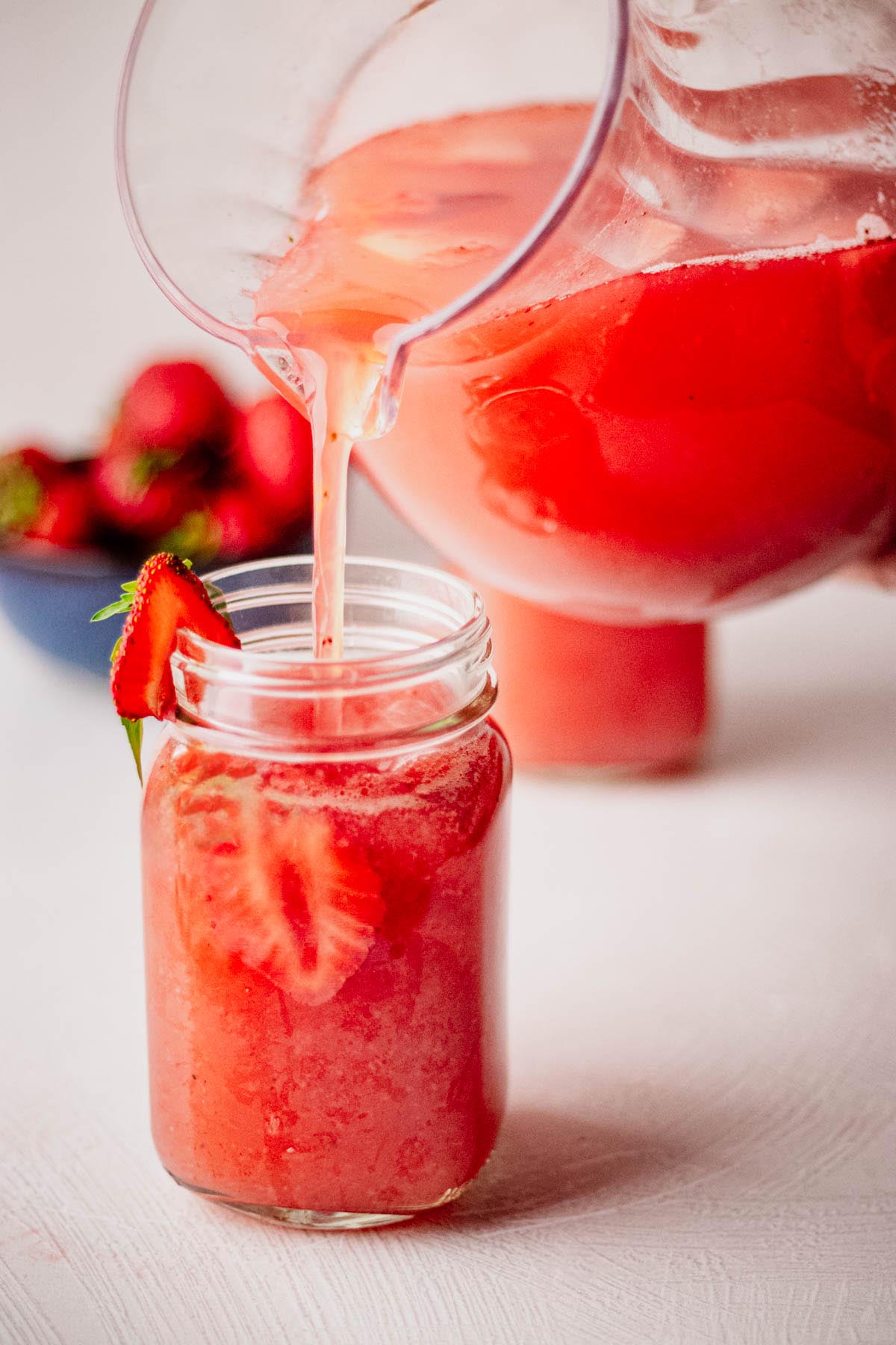 Strawberry agua fresca being poured from a pitcher into a mason jar mug filled with ice and fresh strawberry slices.