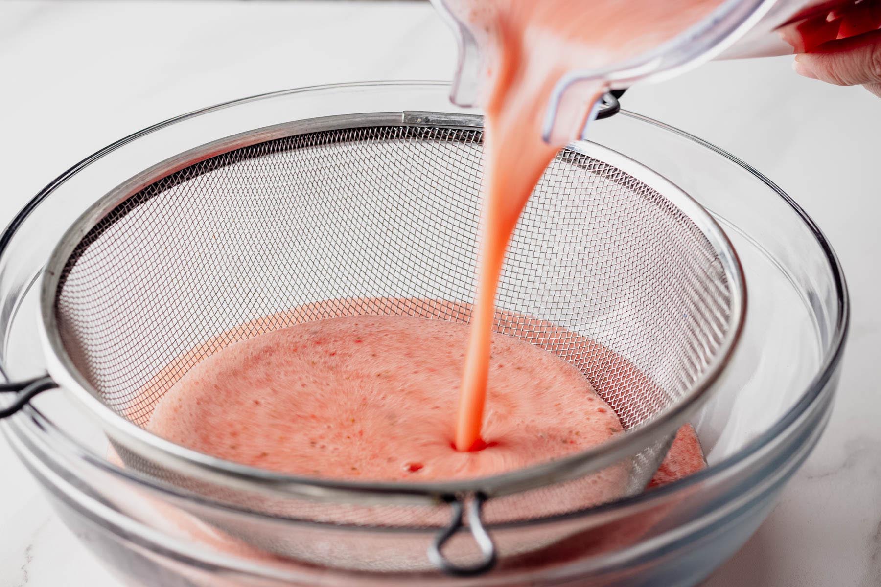 Blended strawberry basil mixture being poured through a fine-mesh strainer into a glass bowl.