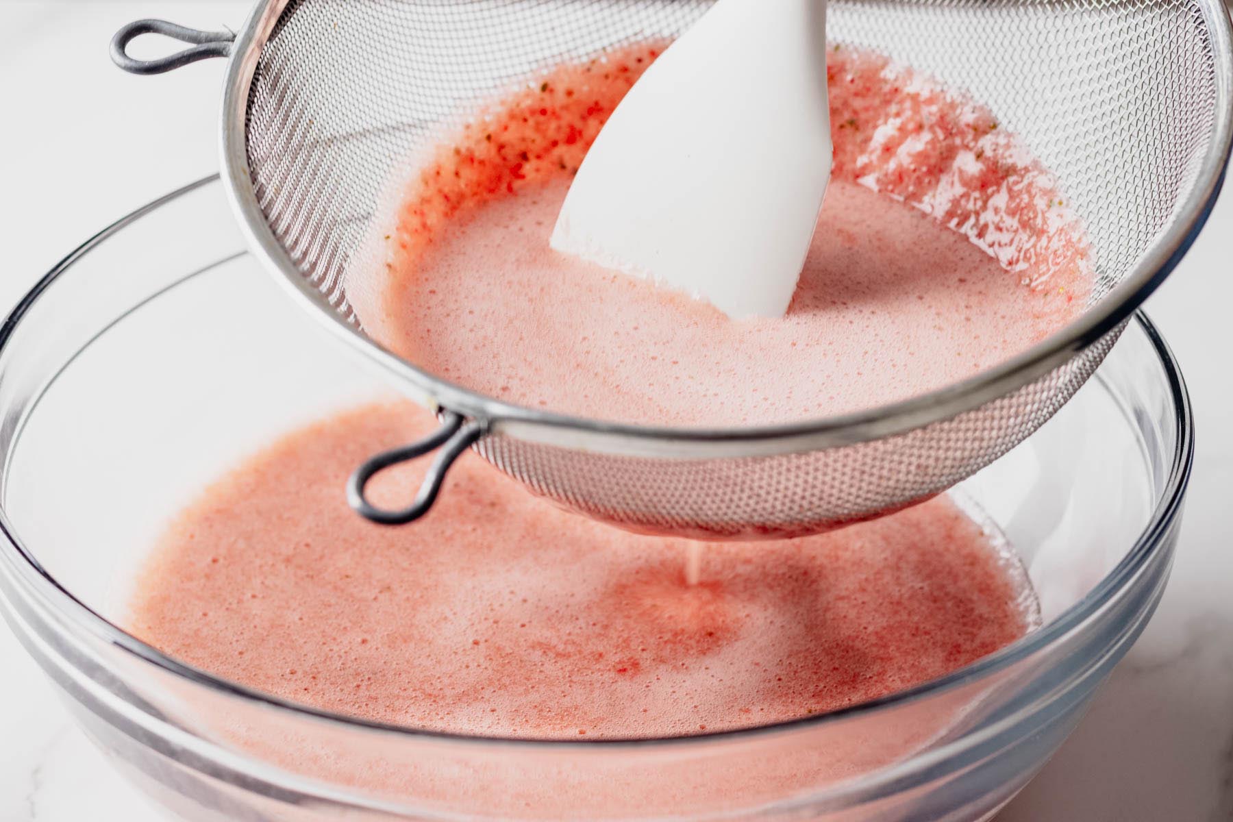 Strawberry basil agua fresca being pressed through a fine-mesh strainer with a spatula to remove seeds and pulp.