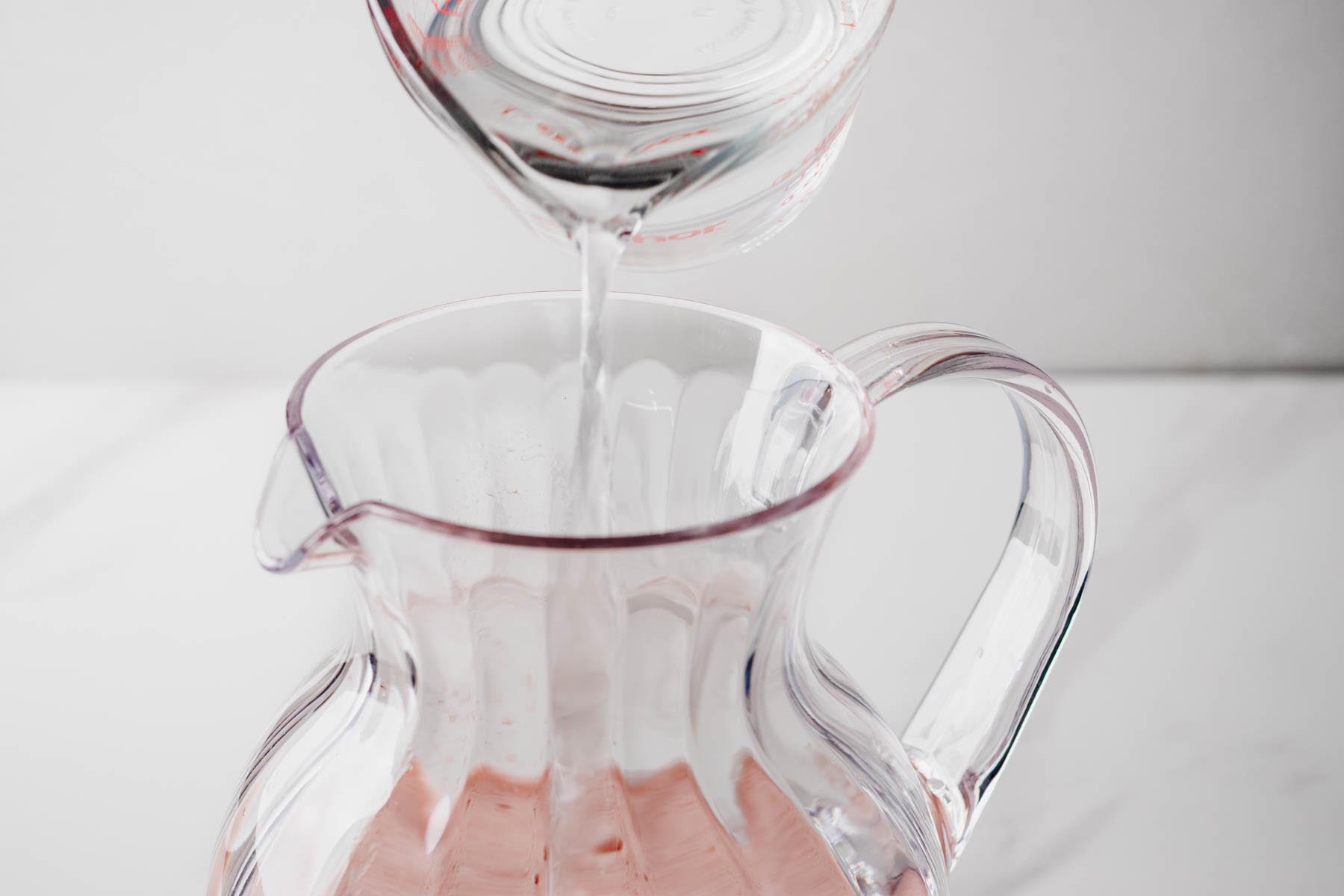 Cold water being added to strained strawberry basil agua fresca in a glass pitcher.