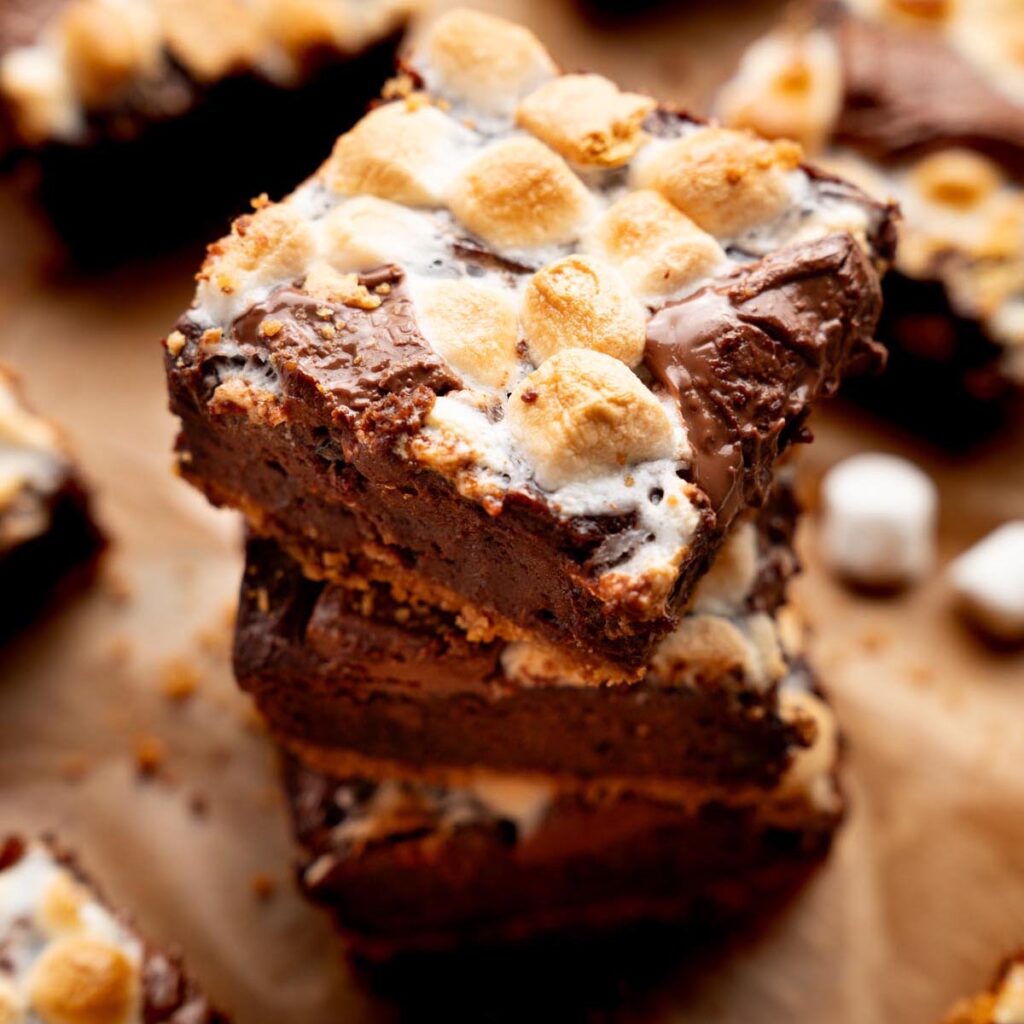 Sweet potato s'mores brownies stacked on a wooden board, topped with toasted mini marshmallows and melted chocolate, with additional brownies blurred in the background.