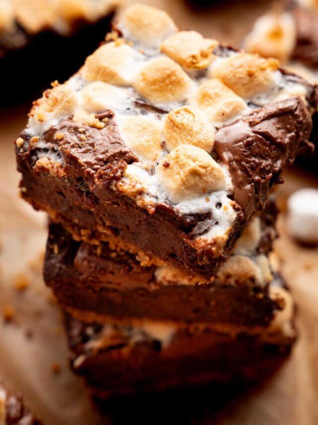 Sweet potato s'mores brownies stacked on a wooden board, topped with toasted mini marshmallows and melted chocolate, with additional brownies blurred in the background.