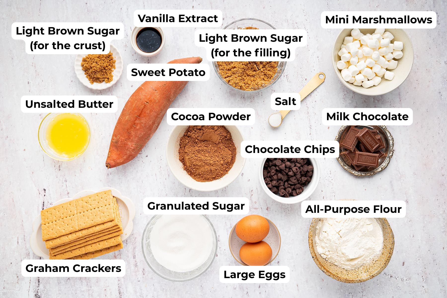 Labeled ingredients for sweet potato s'mores brownies arranged on a white surface, including sweet potato, graham crackers, flour, cocoa powder, eggs, butter, sugars, vanilla extract, chocolate chips, milk chocolate, mini marshmallows, and salt.