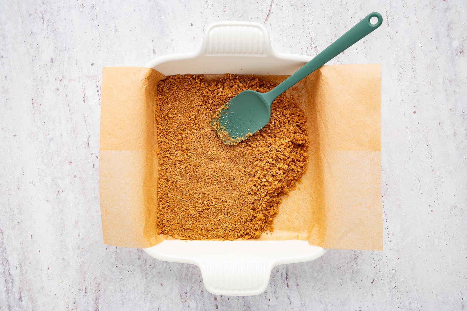 Graham cracker crust mixture pressed firmly into a parchment-lined baking dish.
