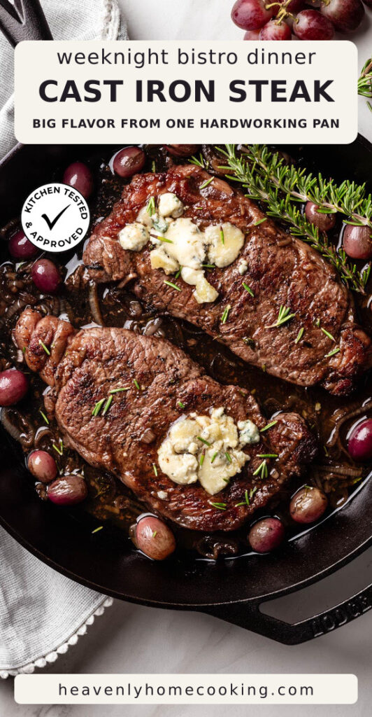 Two cast iron steaks are topped with melting blue cheese and fresh rosemary, surrounded by caramelized onions and red grapes in a cast iron skillet. This vertical Pinterest pin features a homemade cast iron skillet steak recipe from Heavenly Home Cooking.