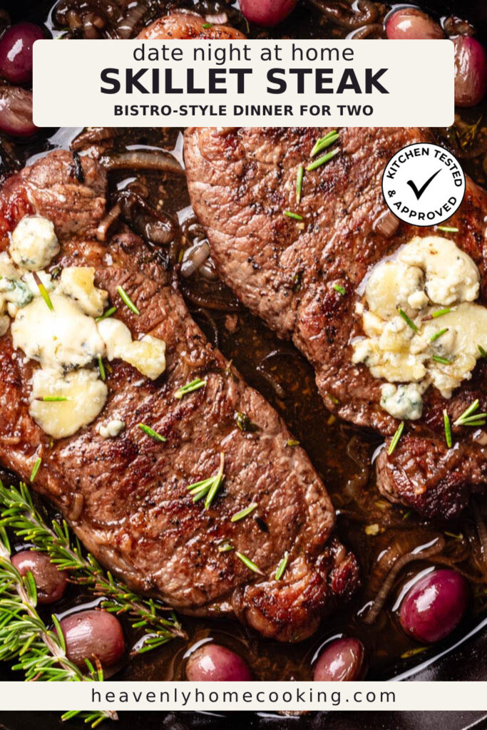 Two cast iron skillet steaks are topped with melting blue cheese and fresh rosemary, surrounded by caramelized onions and roasted red grapes in a rich pan sauce. This vertical Pinterest pin features a homemade cast iron skillet steak recipe from Heavenly Home Cooking.