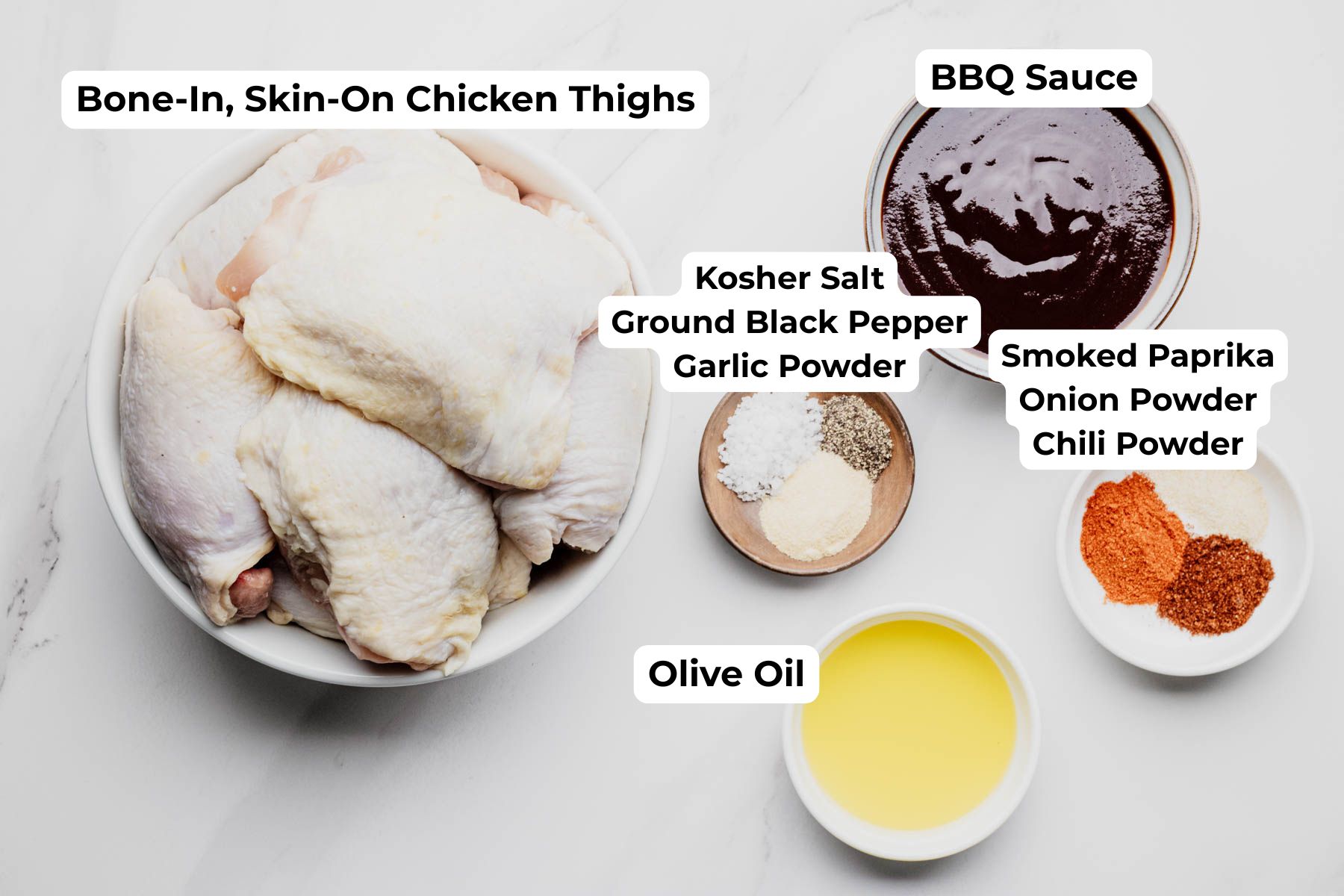 Labeled ingredient photo showing bone-in, skin-on chicken thighs, BBQ sauce, olive oil, kosher salt, ground black pepper, garlic powder, smoked paprika, onion powder, and chili powder for baked BBQ chicken thighs.