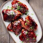 Five baked BBQ chicken thighs coated in a thick, glossy barbecue sauce and garnished with fresh parsley on a gray serving platter.