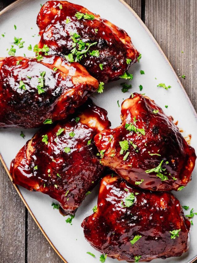 Five baked BBQ chicken thighs coated in a thick, glossy barbecue sauce and garnished with fresh parsley on a gray serving platter.