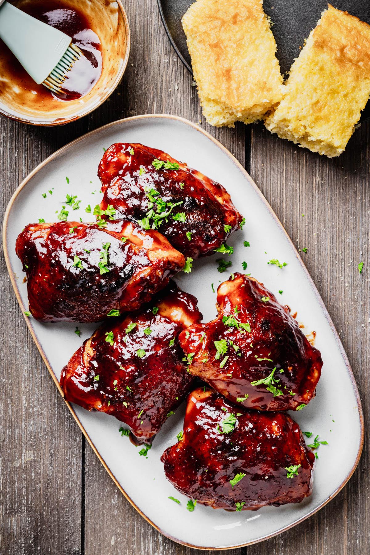 Overhead view of five baked BBQ chicken thighs coated in a thick, glossy barbecue sauce on a serving platter with cornbread and a bowl of barbecue sauce nearby.