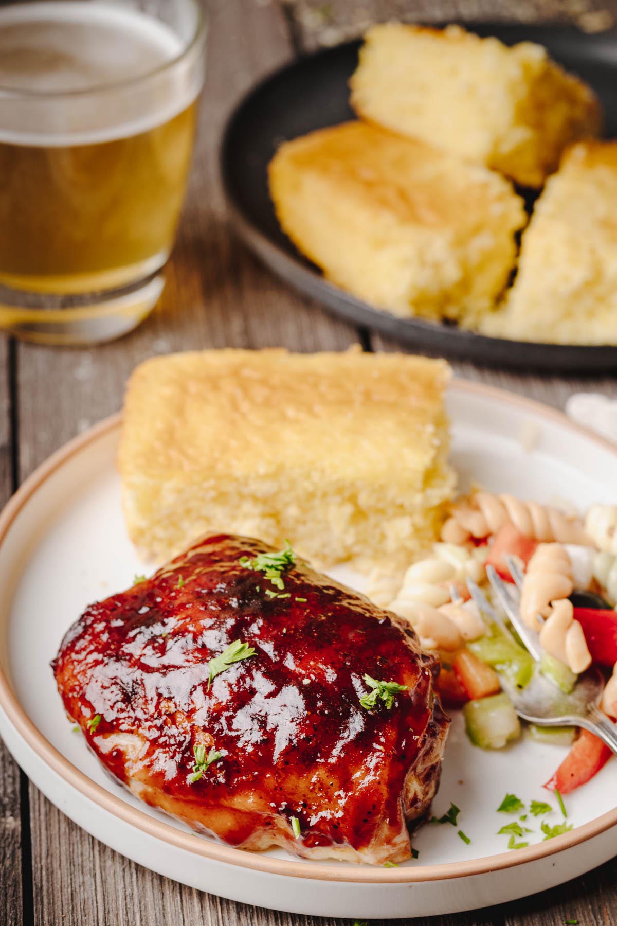 A plated baked BBQ chicken thigh served with cornbread and pasta salad, with a glass of beer and extra cornbread in the background.