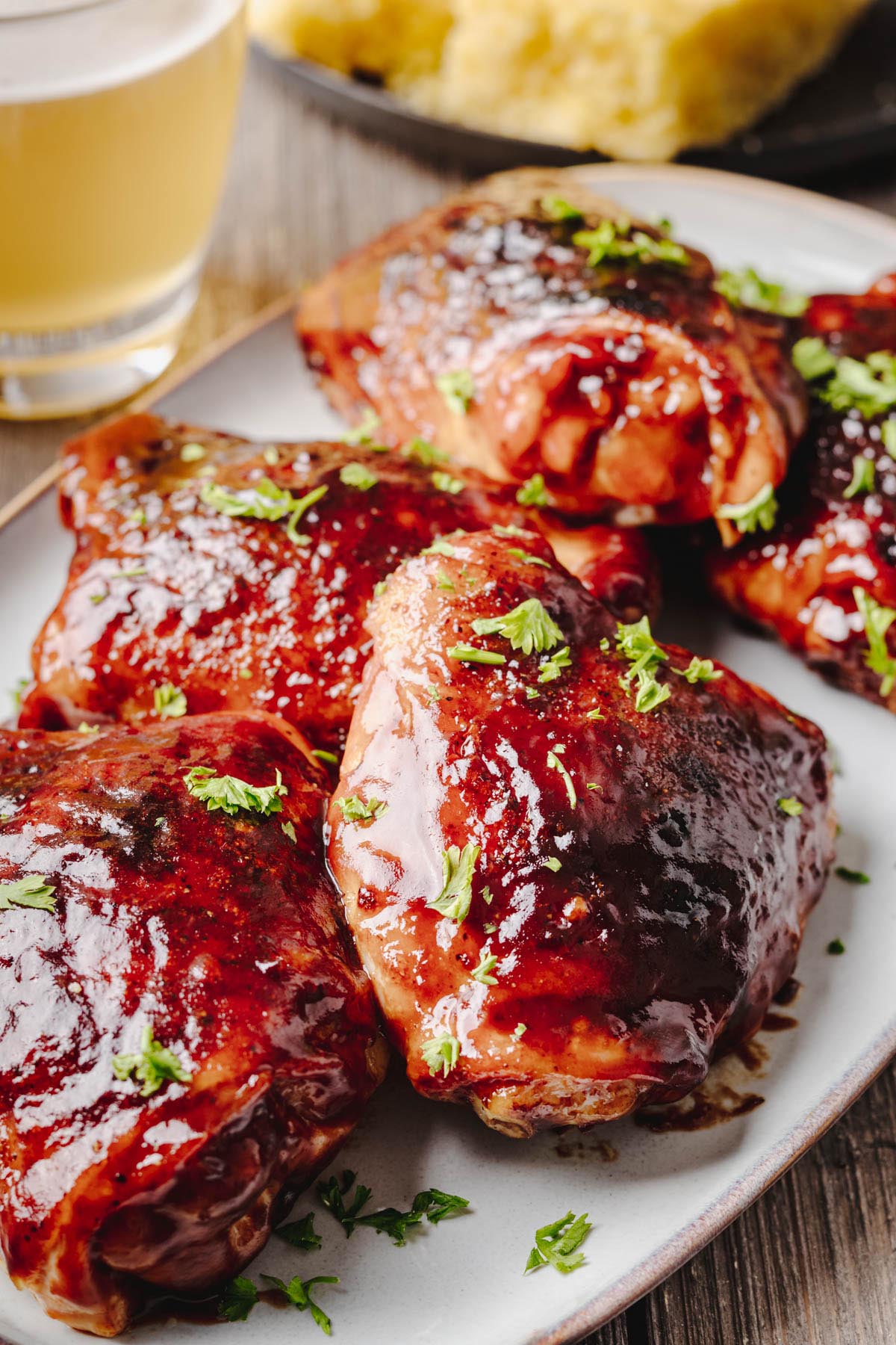 Close-up of baked BBQ chicken thighs coated in a thick, glossy barbecue sauce and garnished with fresh parsley on a serving platter, with cornbread and a glass of beer in the background.