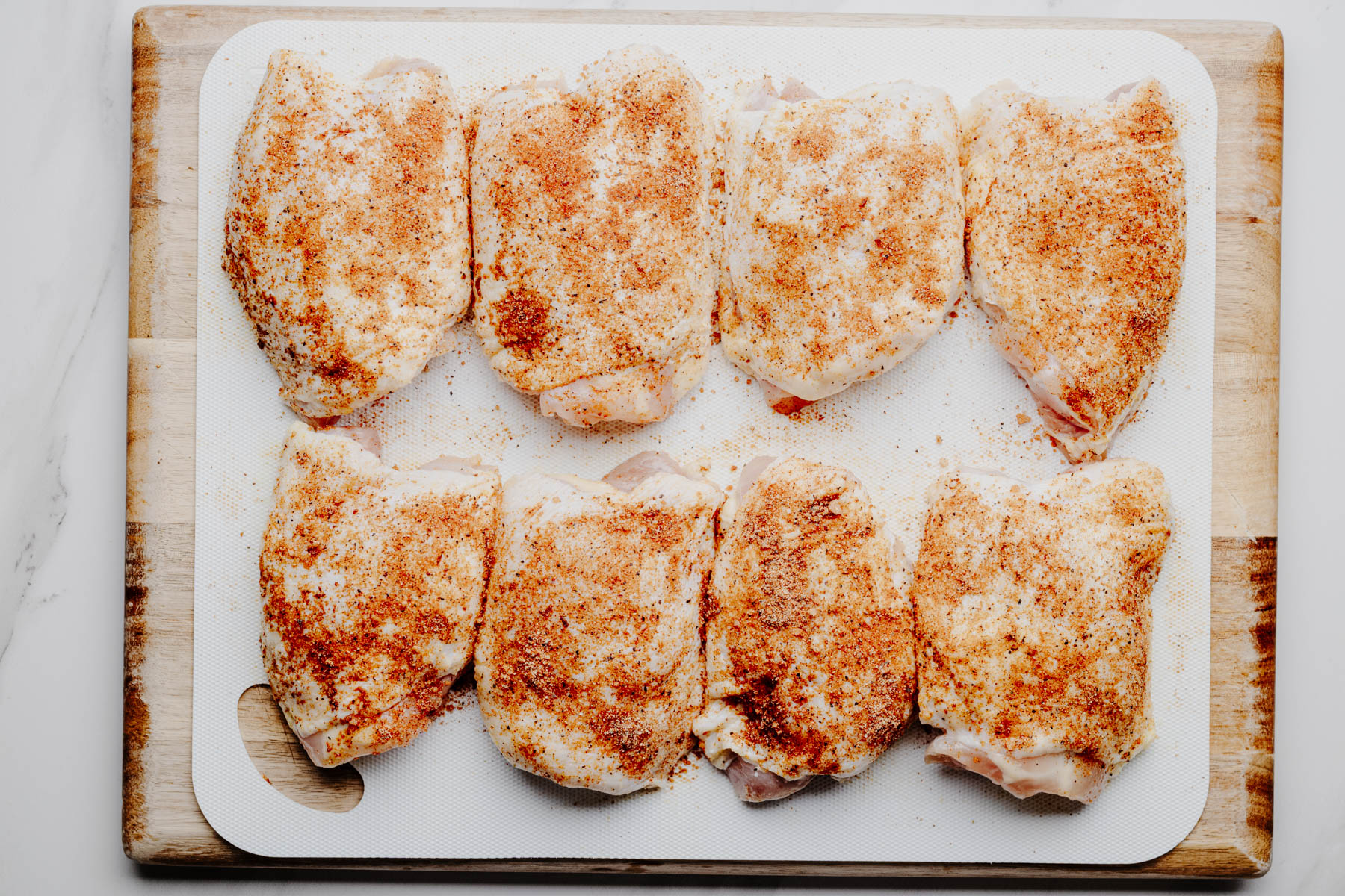 Bone-in, skin-on chicken thighs coated with a homemade spice rub on a white cutting board before baking.