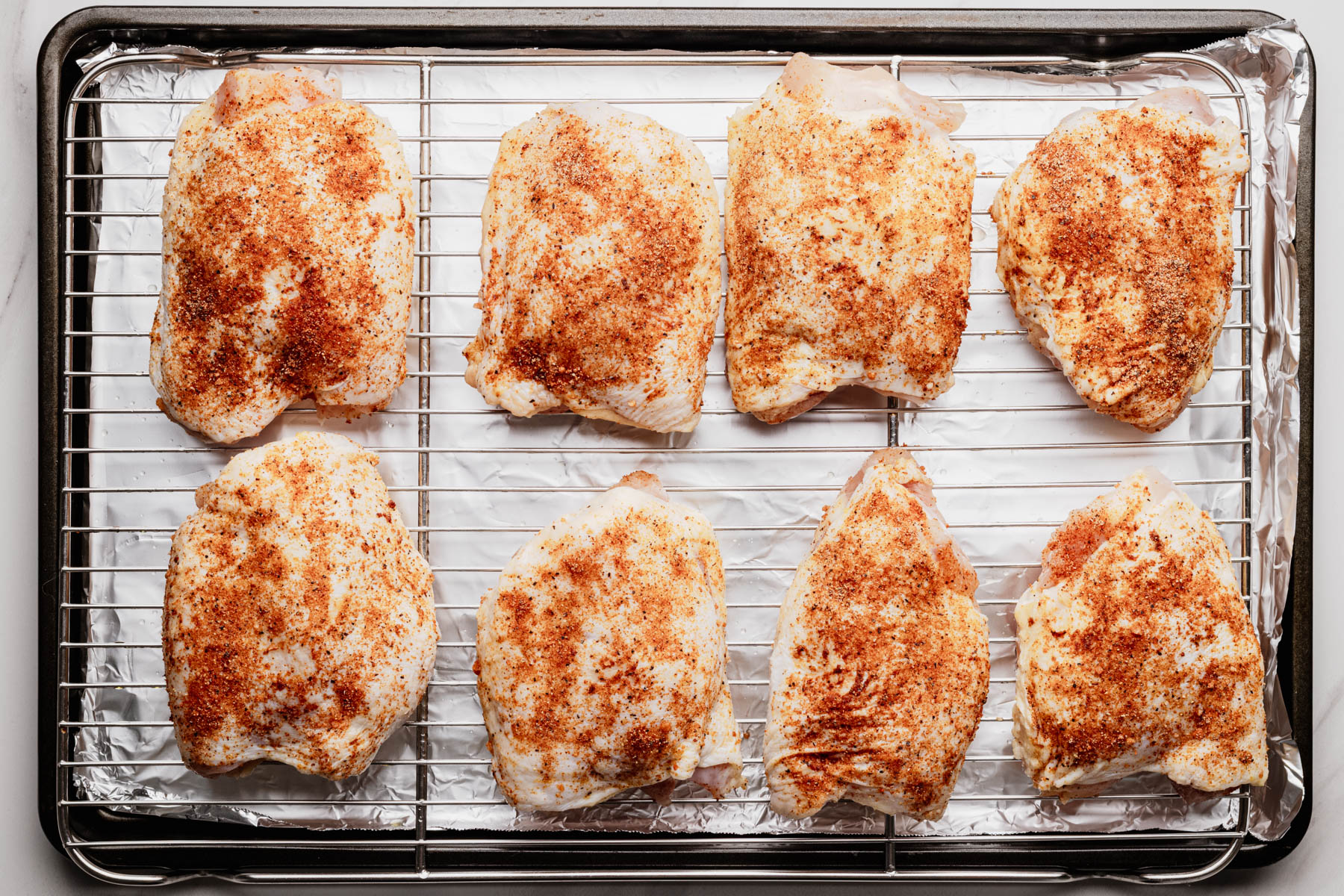 Seasoned bone-in, skin-on chicken thighs arranged on a wire rack over a foil-lined baking sheet before baking.