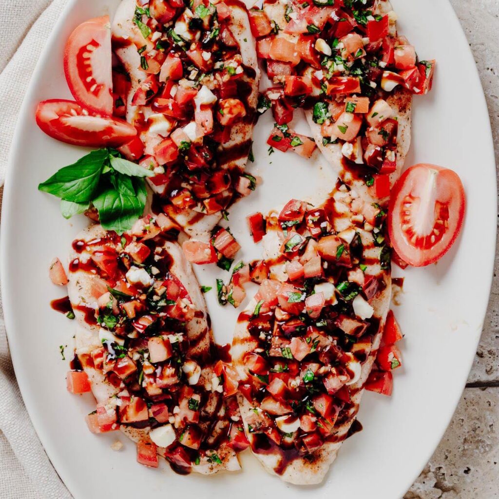 Overhead view of four bruschetta chicken cutlets topped with diced tomatoes, fresh basil, mozzarella pearls, and balsamic glaze on a white platter.