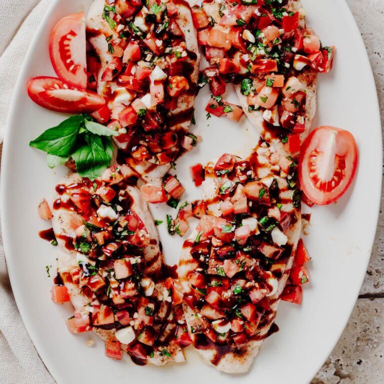 Overhead view of four bruschetta chicken cutlets topped with diced tomatoes, fresh basil, mozzarella pearls, and balsamic glaze on a white platter.