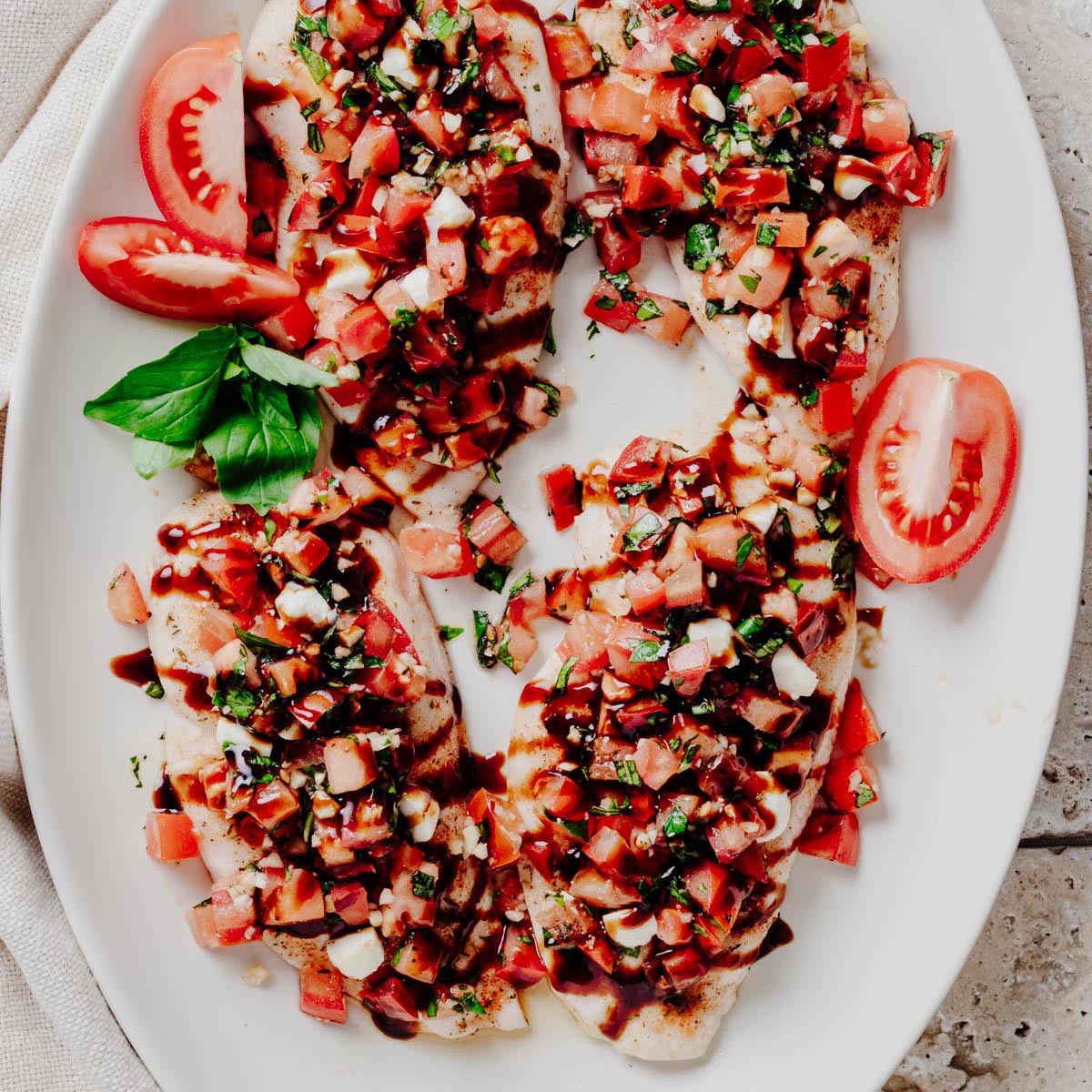 Overhead view of four bruschetta chicken cutlets topped with diced tomatoes, fresh basil, mozzarella pearls, and balsamic glaze on a white platter.