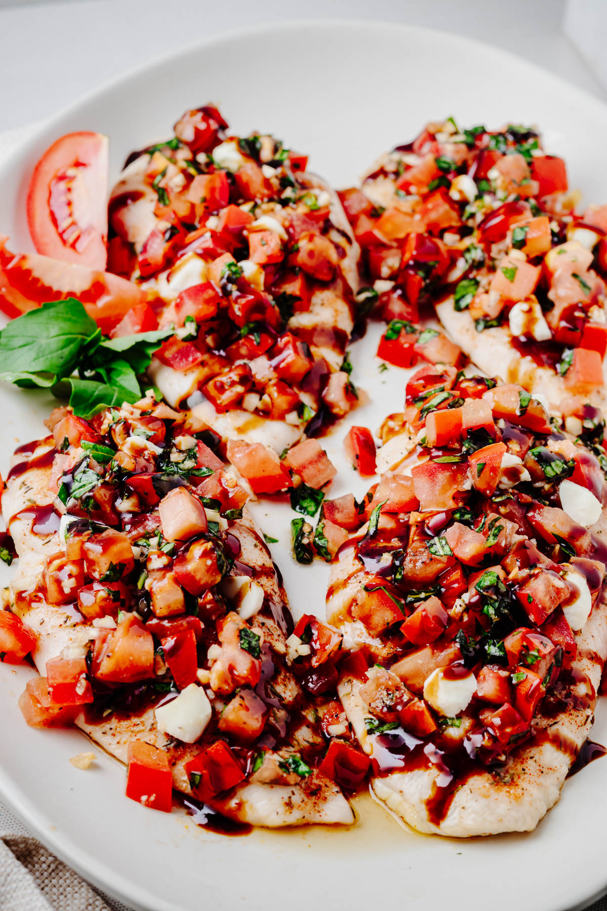 Four baked bruschetta chicken cutlets topped with tomatoes, basil, mozzarella pearls, and balsamic glaze on a white platter.