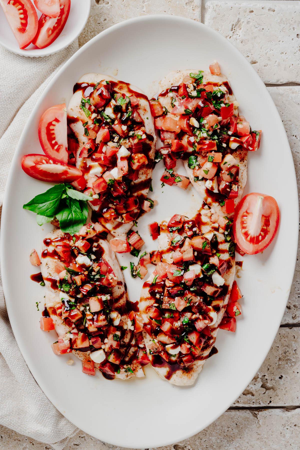 Overhead view of four bruschetta chicken cutlets on a white platter with fresh tomatoes and basil.