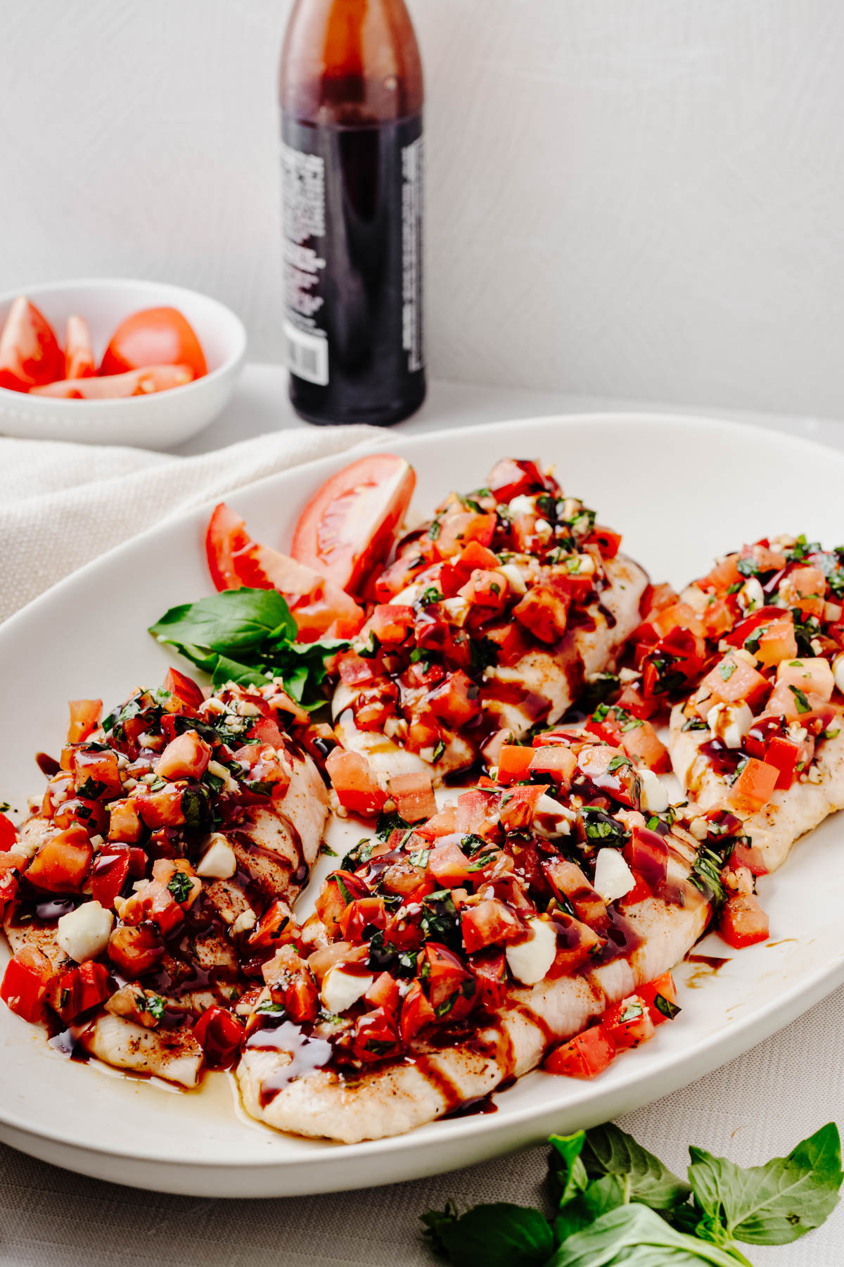 Bruschetta chicken topped with tomato basil bruschetta, mozzarella pearls, and balsamic glaze, with fresh basil and tomatoes nearby.