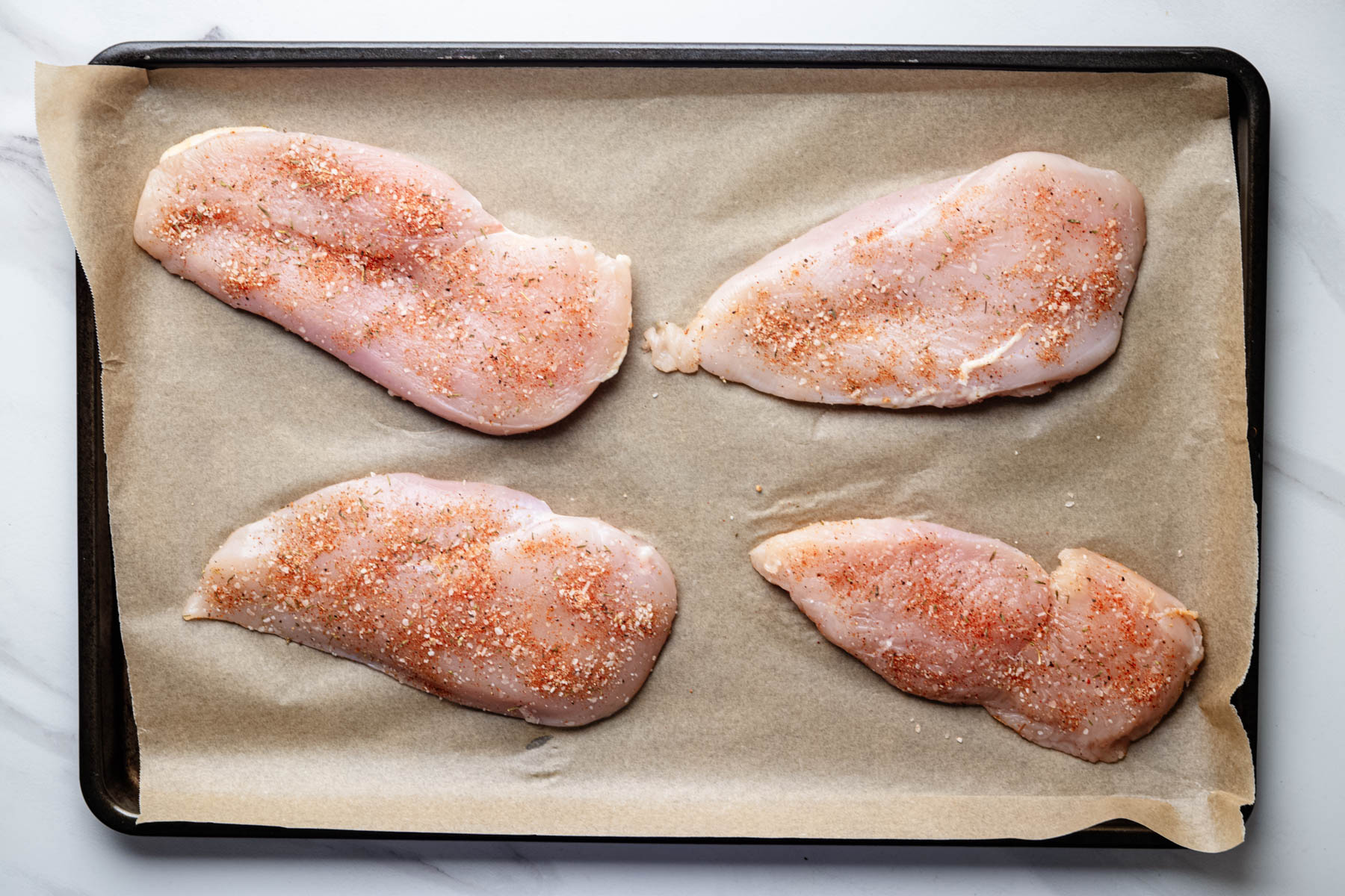 Raw chicken breast cutlets seasoned with garlic herb dry brine on a parchment-lined baking sheet.