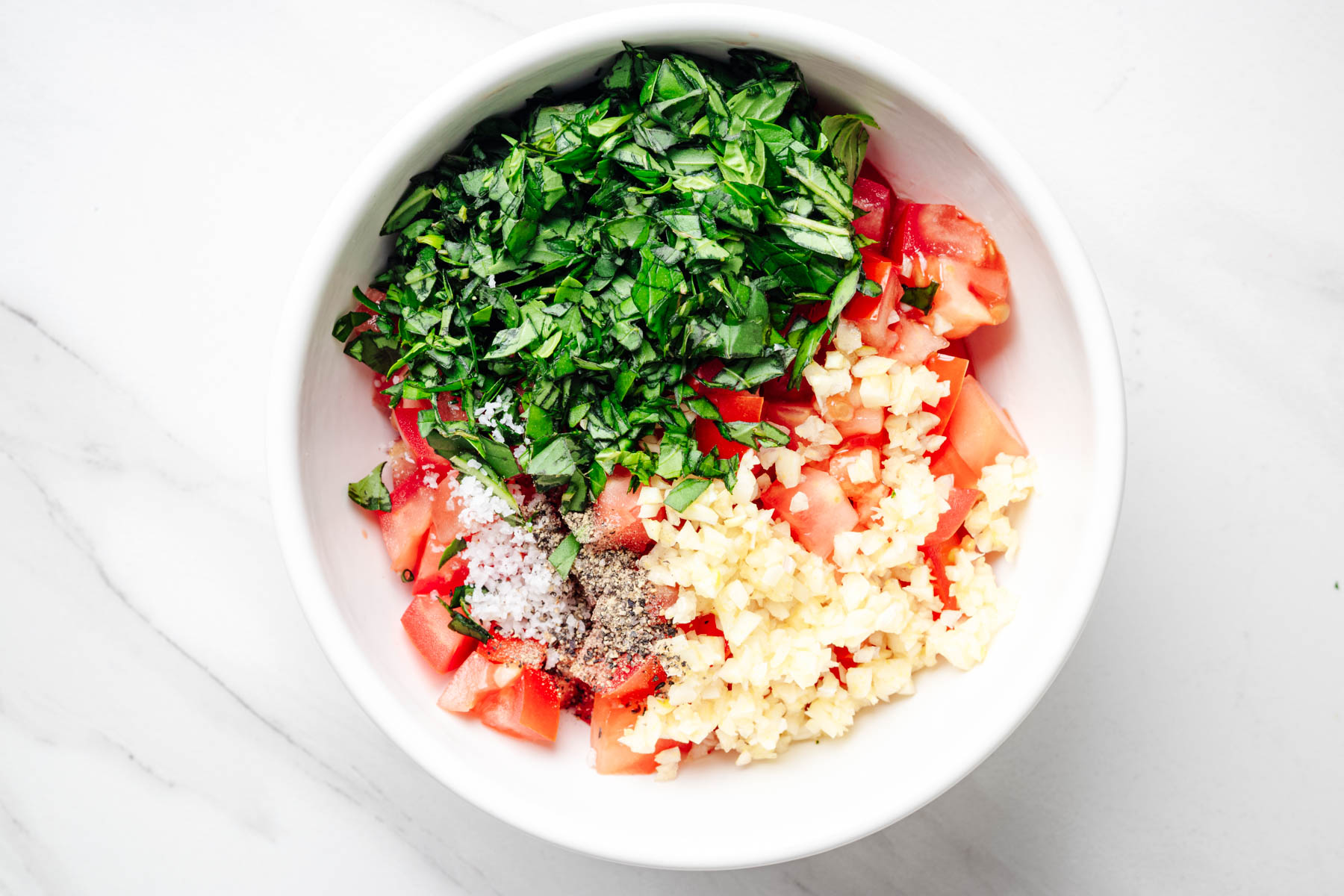 Diced Roma tomatoes, chopped fresh basil, minced garlic, salt, and black pepper in a white mixing bowl.