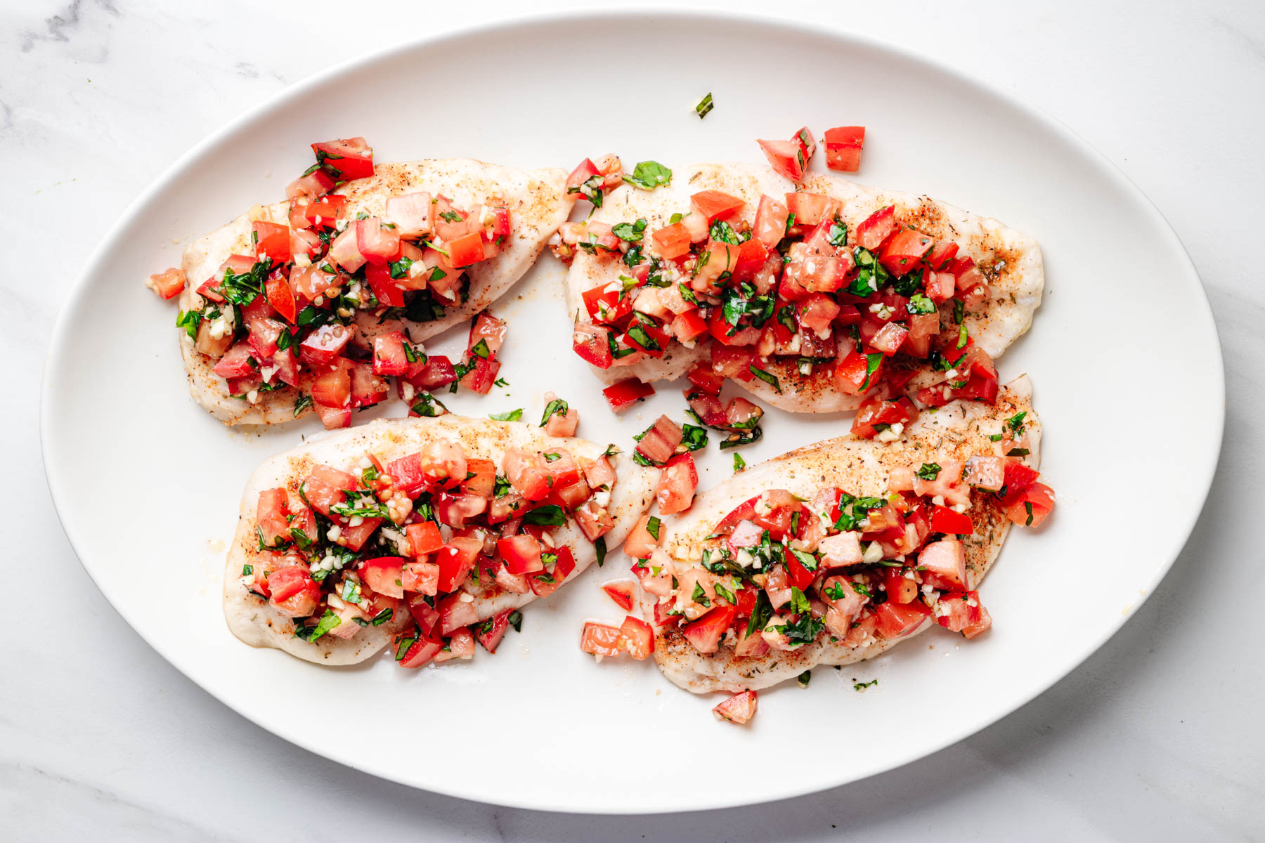 Baked chicken breast cutlets topped with fresh tomato, basil, and garlic bruschetta on a white platter.