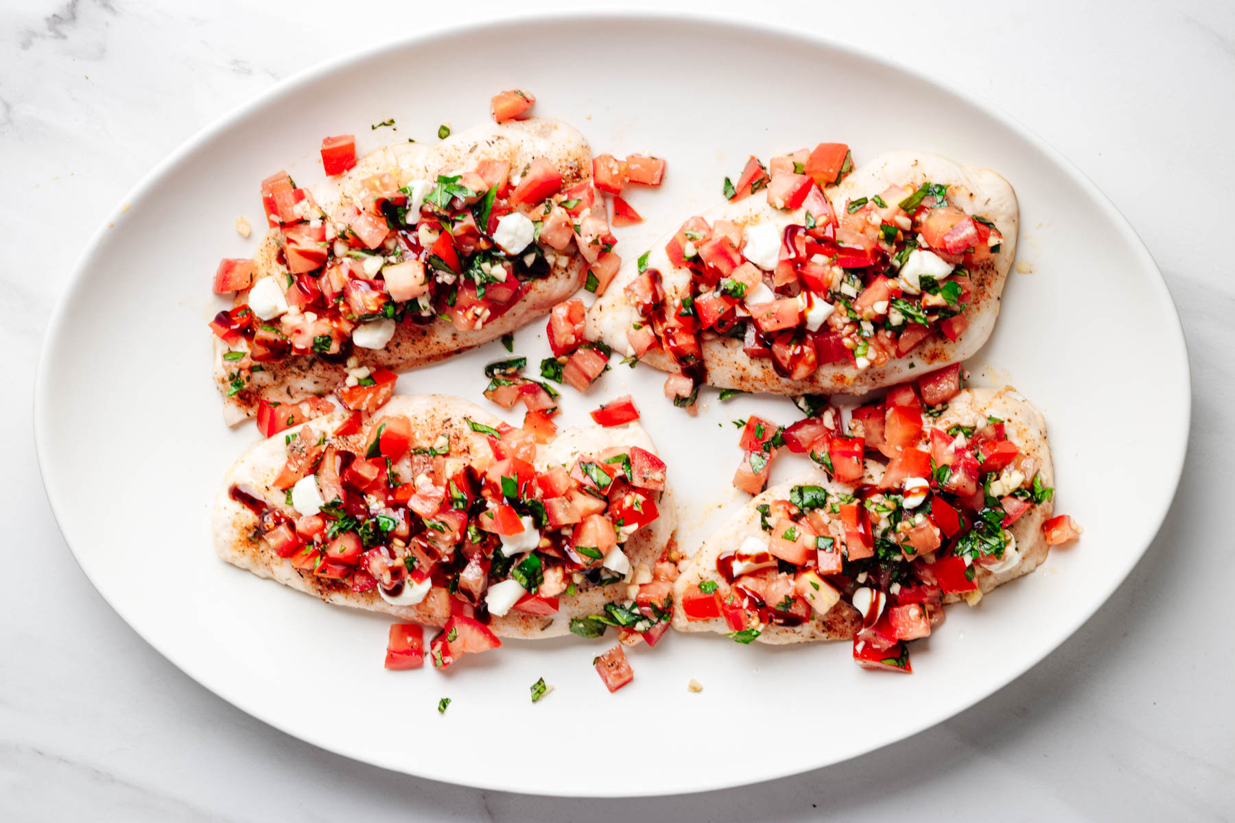 Bruschetta chicken topped with mozzarella pearls and drizzled with balsamic glaze on a white serving platter.