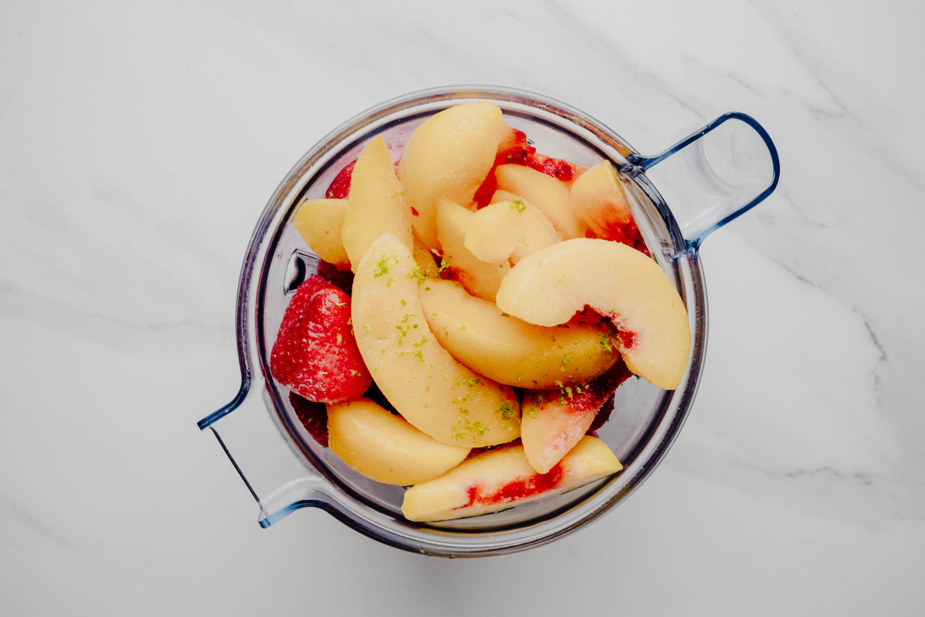 Overhead view of frozen peaches, frozen strawberries, watermelon, lime zest, and other cold fruit soup ingredients in a blender.