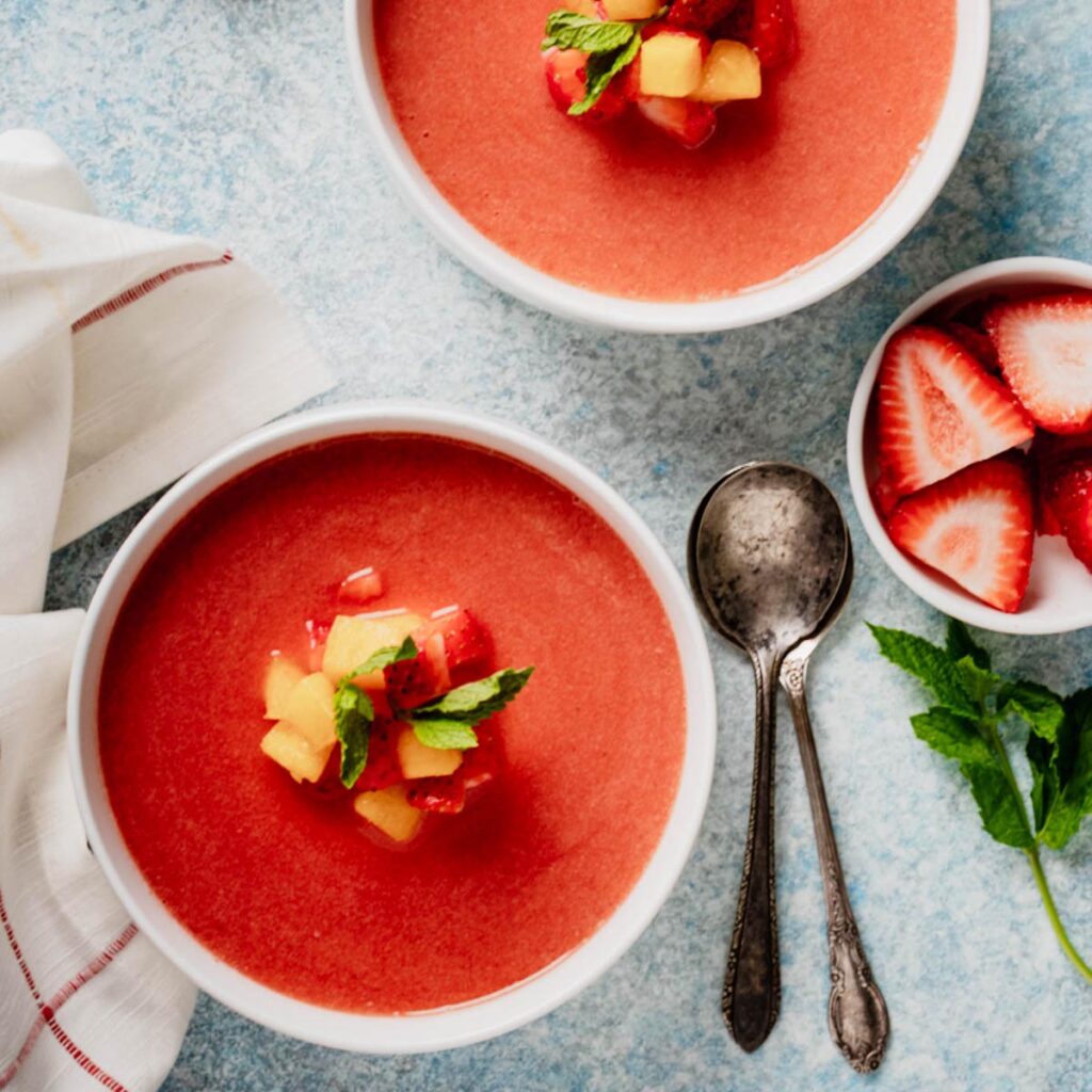Two bowls of cold fruit soup garnished with peaches, strawberries, and mint, surrounded by fresh fruit and antique spoons.