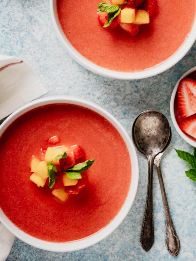 Two bowls of cold fruit soup garnished with peaches, strawberries, and mint, surrounded by fresh fruit and antique spoons.