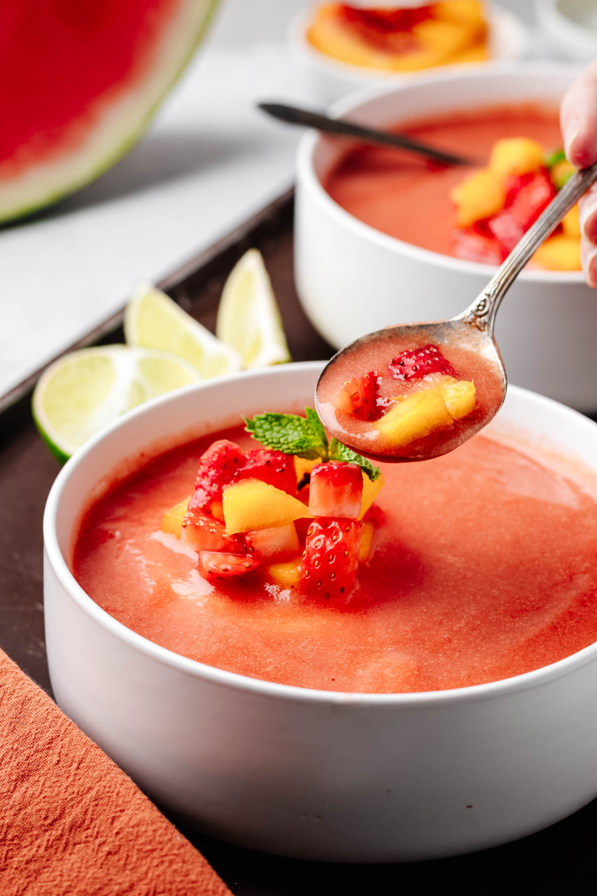 A spoonful of cold fruit soup with diced peaches and strawberries lifted above a garnished bowl.