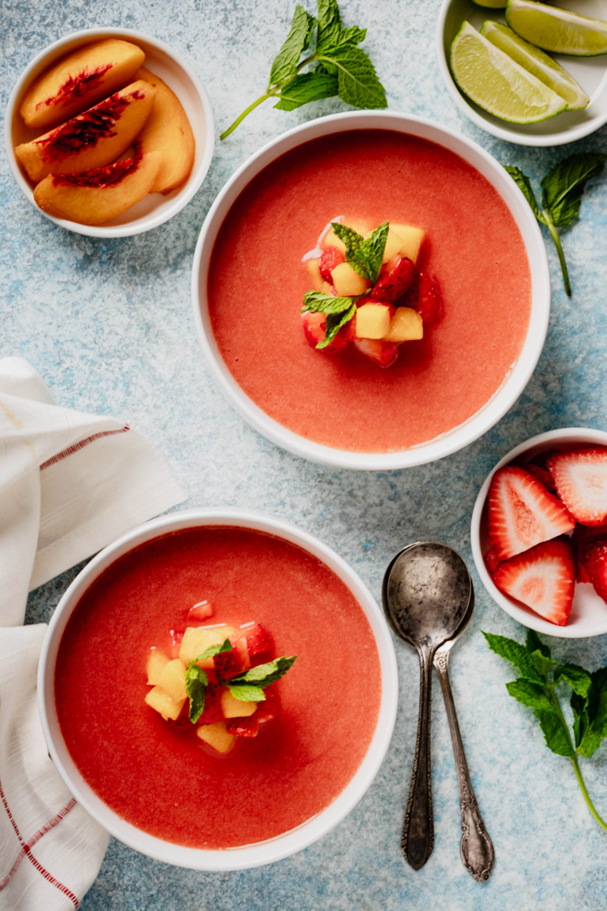 Overhead view of two bowls of cold fruit soup with peaches, strawberries, lime wedges, mint, and spoons.