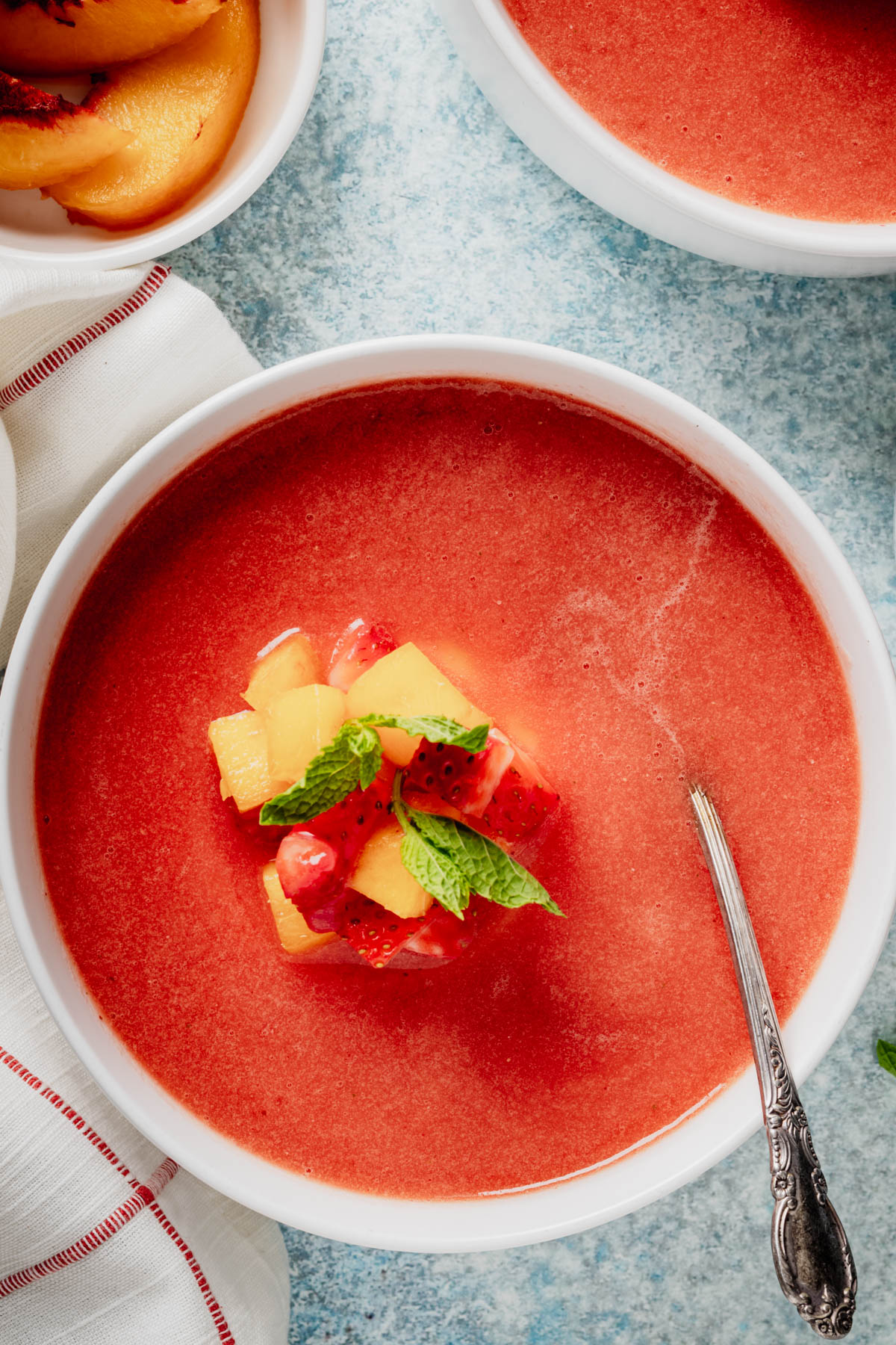 Overhead view of cold fruit soup topped with diced peaches, strawberries, and mint, with a spoon resting in the bowl.