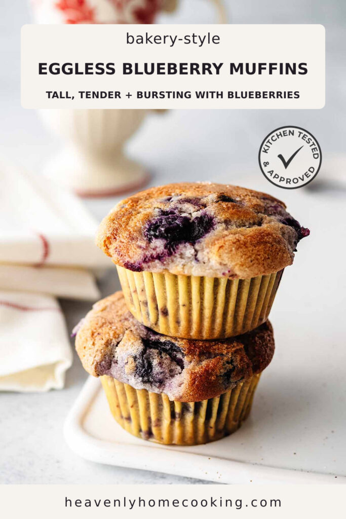 Two bakery-style eggless blueberry muffins are stacked on a marble board, showing tall domed tops bursting with juicy blueberries. The Pinterest pin includes the text "Bakery-Style Eggless Blueberry Muffins – Tall, Tender + Bursting with Blueberries." This vertical Pinterest pin features a homemade eggless blueberry muffins recipe from Heavenly Home Cooking.