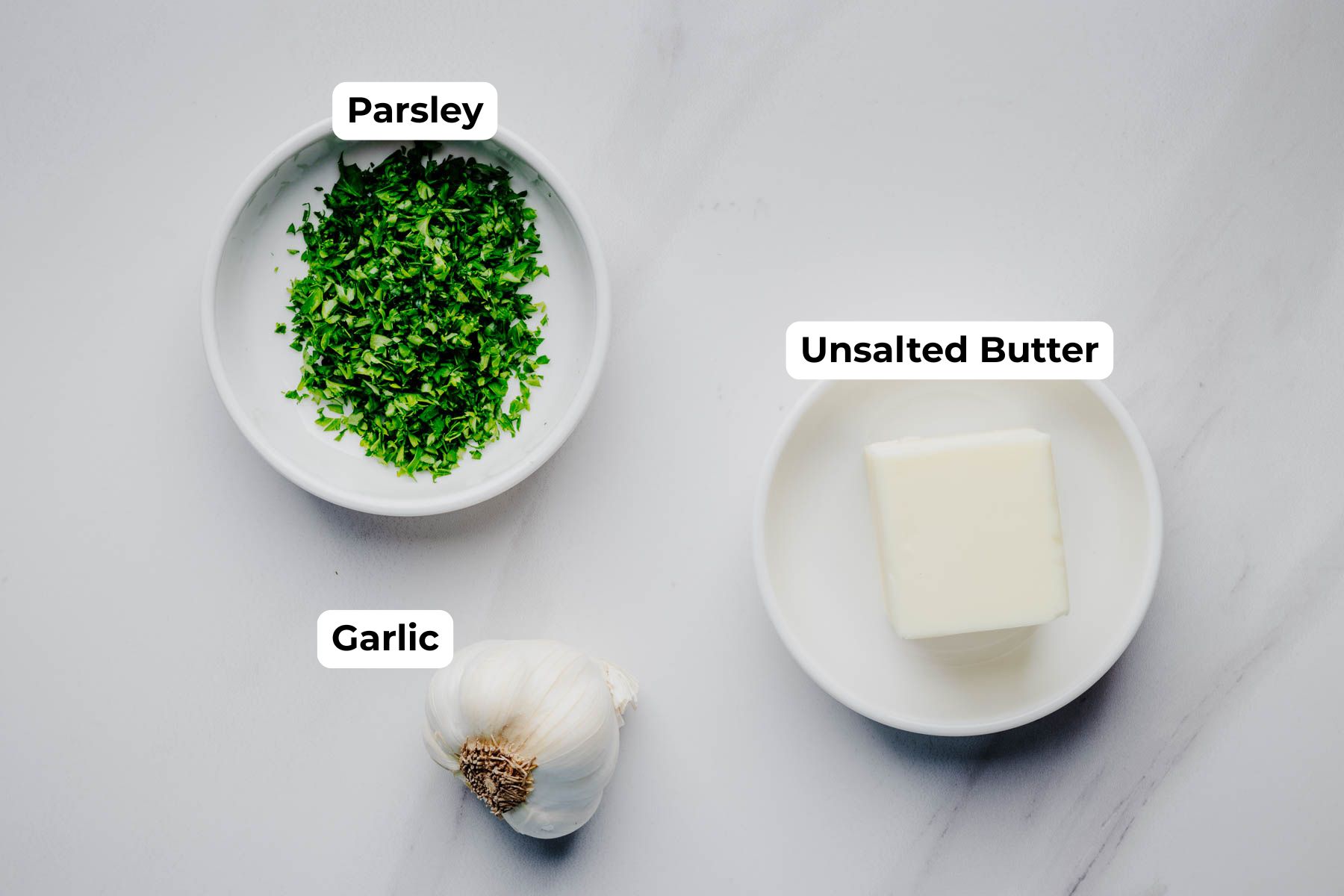 Overhead view of the labeled garlic butter topping ingredients for garlic dinner rolls, including chopped parsley, fresh garlic, and unsalted butter.